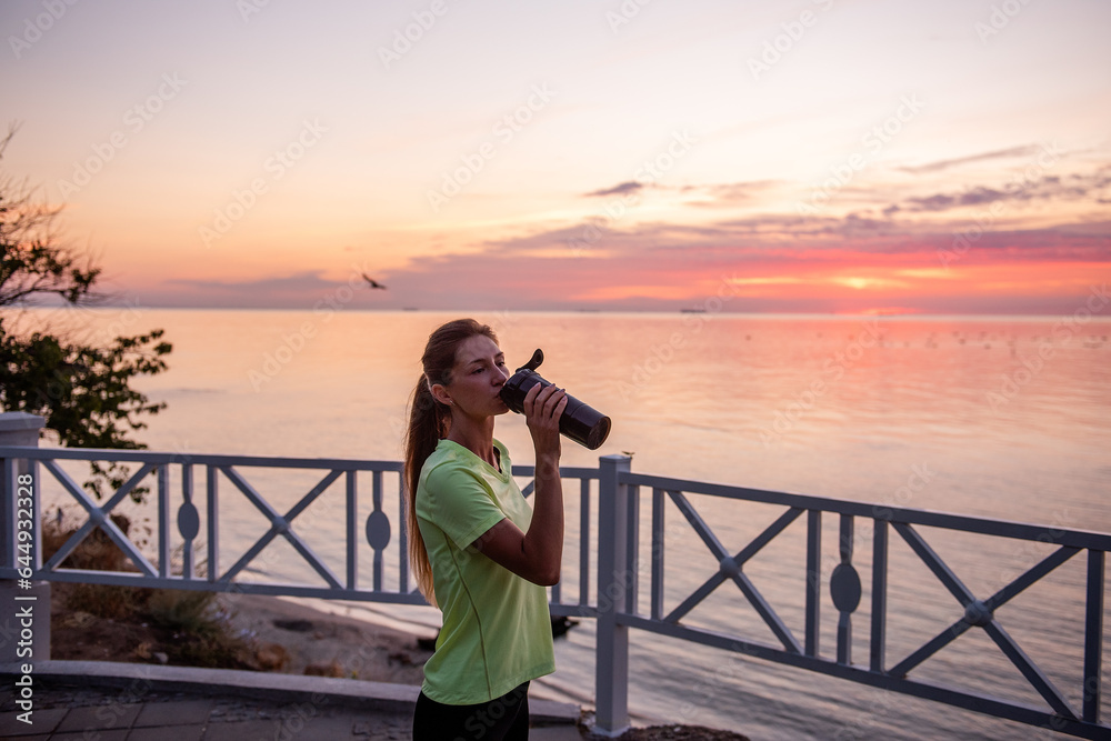 Obraz premium Portrait of an athlete who drinks water from a sports bottle. A young woman quenches her thirst with a fitness drinker. Maintaining water balance during training at sunrise. Girl's morning routine.