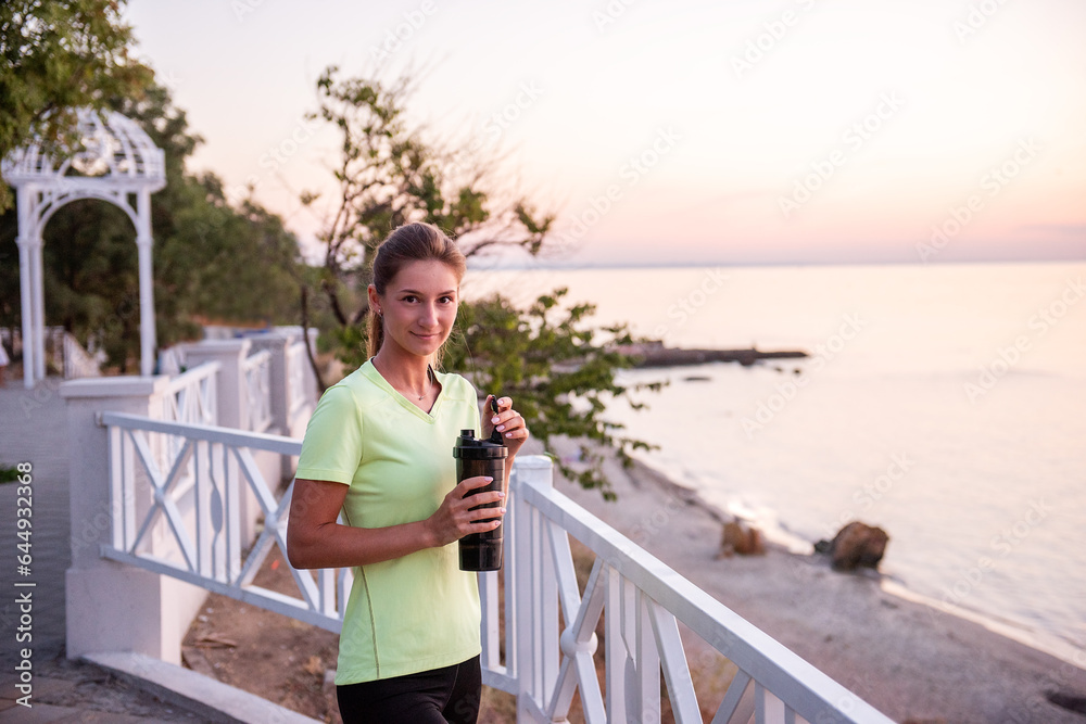 Portrait of an athlete who drinks water from a sports bottle. A young woman quenches her thirst with a fitness drinker. Maintaining water balance during training at sunrise. Girl's morning routine. Portrait of an athlete who drinks water from a sports bottle. A young woman quenches her thirst with a fitness drinker. Maintaining water balance during training at sunrise. Girl's morning routine.