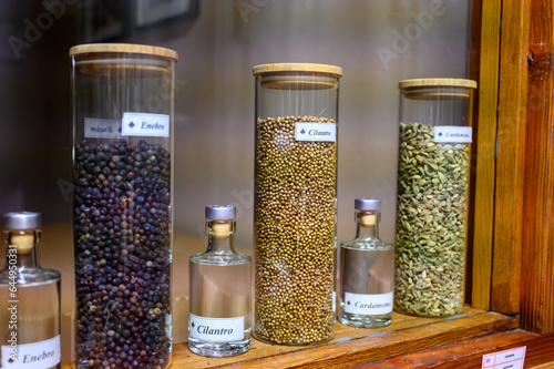 Tastes of gin, botanicals ingredients for gin distillery process, name in Spaniish translated into English: juniper, cardamom and coriander, dried coriander seeds in glass pot