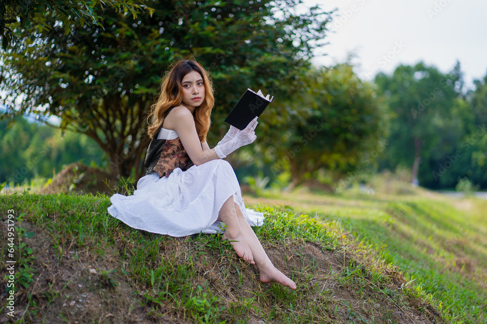 Fototapeta premium A portrait of a beautiful girl sitting on a green meadow under tree branches with a book, a woman reading on nature, concept hobbies, and lifestyle, Read, book, Novel
