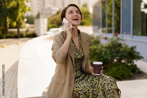 Outdoor photo of stylish beautiful woman with short haircut  enjoing autu,n day  holding cup of coffee, using mobyle phone. Weaering casual  jacker and elegant dress. photo