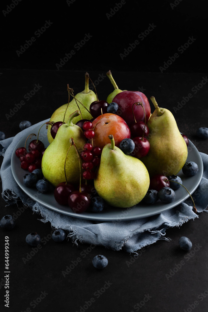 Against a dark background, a still life of pears, plums, red currants, blueberries and cherries in a gray plate. Against a dark background, a still life of pears, plums, red currants, blueberries and cherries in a gray plate.