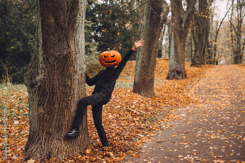 Halloween. Pumpkinhead. A man with a pumpkin on his head stands in a park on an alley strewn with yellow autumn leaves.