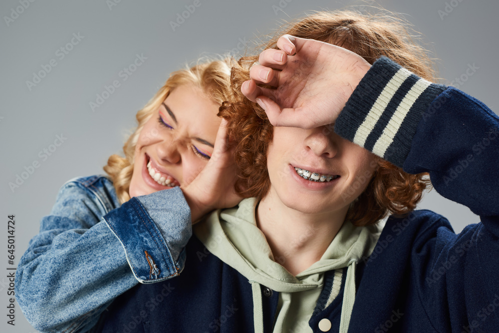 cheerful blonde teenage girl with closed eyes obscuring face of stylish redhead friend on grey cheerful blonde teenage girl with closed eyes obscuring face of stylish redhead friend on grey