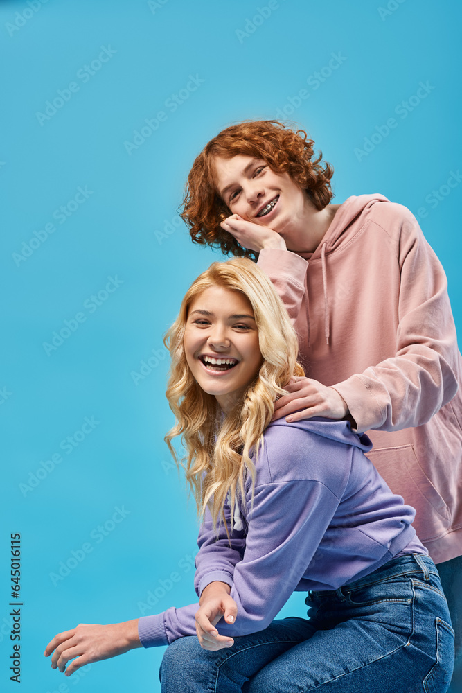 happy redhead teenage boy leaning on blonde girlfriend looking at camera and laughing on blue happy redhead teenage boy leaning on blonde girlfriend looking at camera and laughing on blue