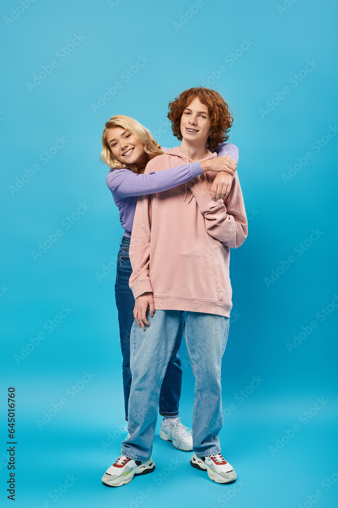 joyful blonde teen girl embracing redhead boyfriend, happy friends in hoodies and jeans on blue joyful blonde teen girl embracing redhead boyfriend, happy friends in hoodies and jeans on blue