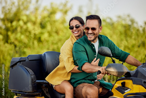 A close-up of a smiling love couple enjoys the quad bike ride and looks at the nature around them.