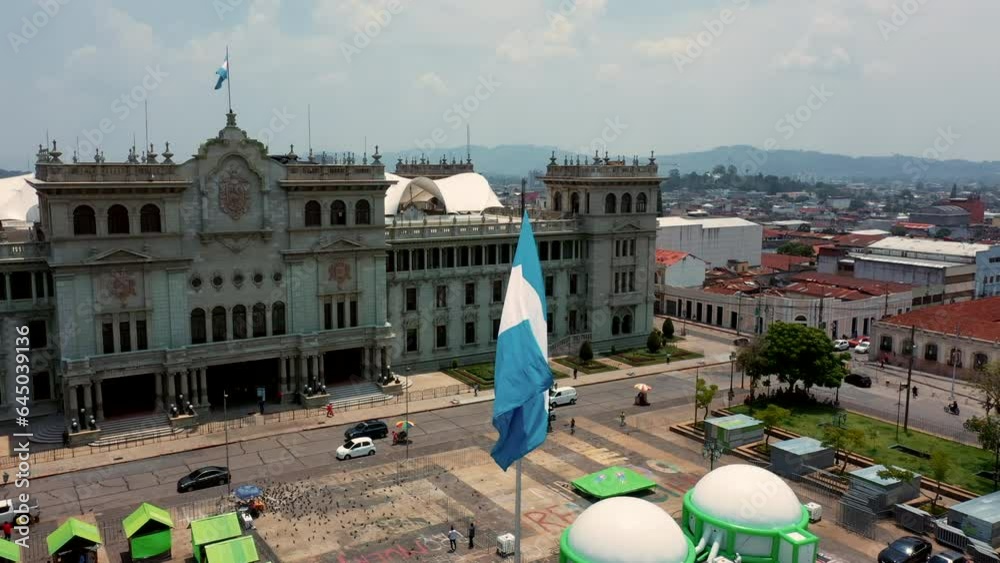 Bandera Guatemalteca ondenado al viento sobre asta en plaza municipal de la ciudad con palacio nacional de cultura de fondo  guatemala y la catedral metropolitana en el centro de la ciudad