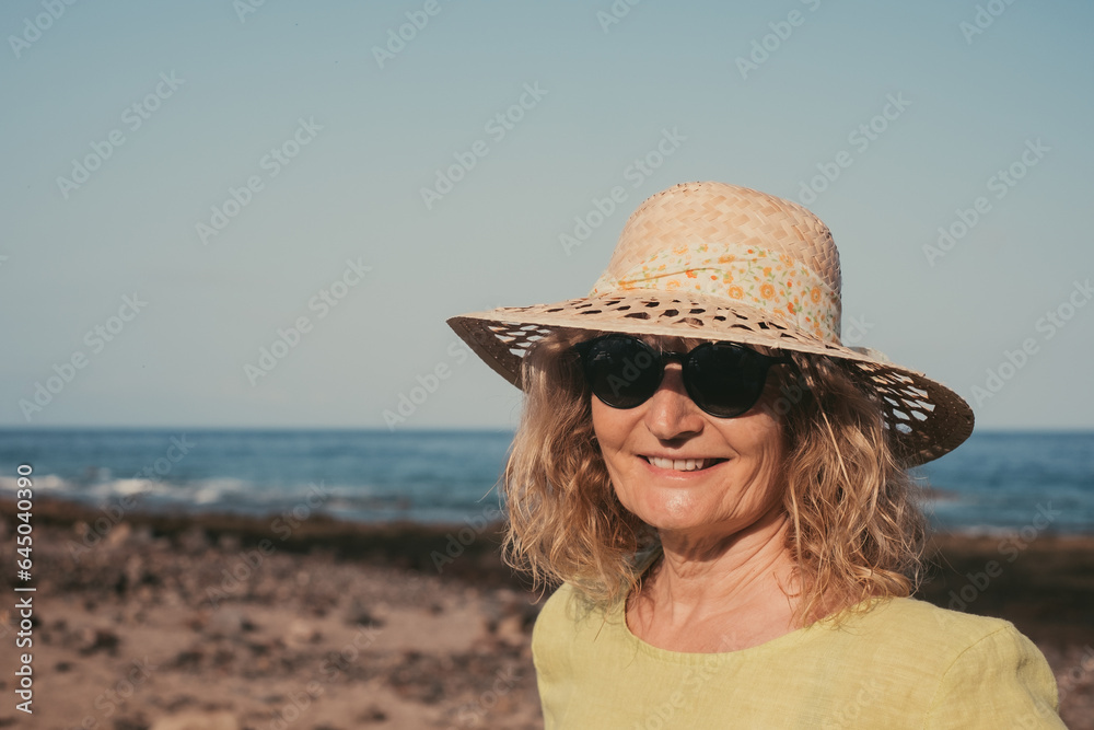 Portrait of happy middle aged blonde woman standing outdoors at the sea beach wearing sunglasses and hat. Vacation, freedom, concept. Horizon over water