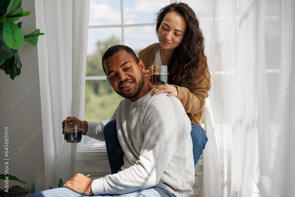 Smiling interracial couple holding cups of coffee while sitting and relaxing near window in morning Smiling interracial couple holding cups of coffee while sitting and relaxing near window in morning