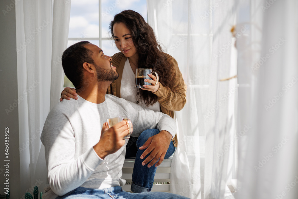 Obraz premium Smiling young asian woman hugging african american boyfriend with coffee on windowsill at home in morning