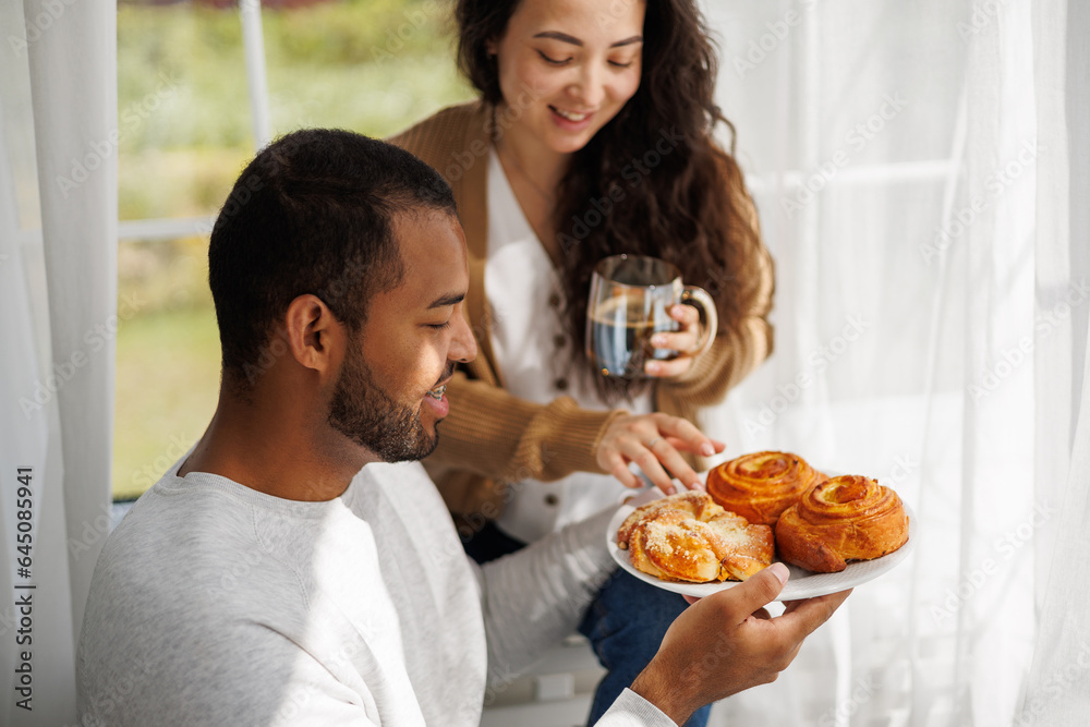 Smiling young african american man holding homemade pastry near asian girlfriend with coffee at home Smiling young african american man holding homemade pastry near asian girlfriend with coffee at home