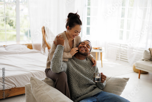 Smiling asian woman hugging african american boyfriend in sweater with coffee on armchair at home