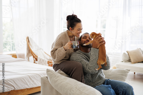 Smiling asian woman in sweater holding coffee near african american boyfriend playing with dog at home