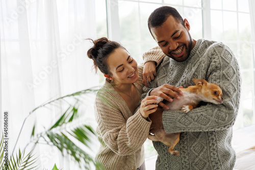 Smiling young diverse couple in cozy clothes petting chihuahua dog at home