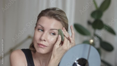 Middle aged beautiful woman making herself facial massage with gua sha behind the mirrow at home photo