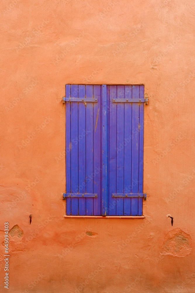 Colourful old-fashioned window of blue colour on the vivid orange weathered background. Closed vibrant shutters on simple worn wall in France