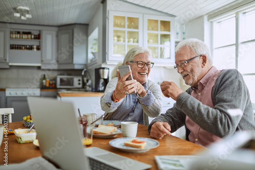 Senior Caucasian couple using a smartphone while having breakfast together in the kitchen in the morning photo