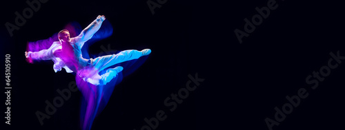 Young guy in stylish white clothes in motion, jumping on black studio background in neon with mixed lights effect. Concept of movements, art, dance and sport, fashion, youth. Banner. Copy space for ad