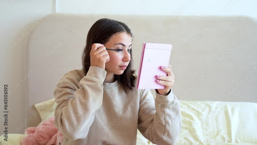 Teenage girl putting makeup. Young woman looking at mirror and apply mascara on eyelashes in bedroom. Cosmetics for youth culture trend. Daily make-up. Copy space Teenage girl putting makeup. Young woman looking at mirror and apply mascara on eyelashes in bedroom. Cosmetics for youth culture trend. Daily make-up. Copy space