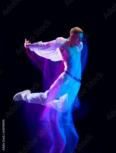 Fototapeta Naklejka Na Ścianę i Meble -  Muscular young man in white stylish clothes jumping against black studio background in neon with mixed lights effect. Concept of movements, art, dance and sport, fashion, youth, ad