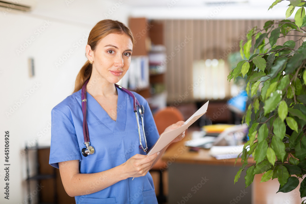 Young female therapist studies important medical documents while standing in the clinic's office - obrazy, fototapety, plakaty 