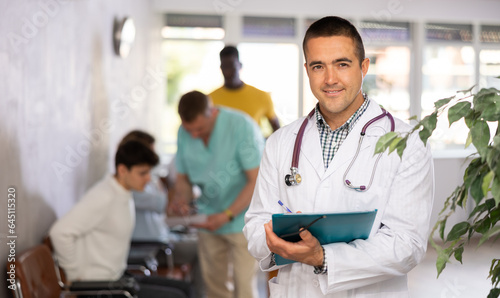 Portrait of smiling polite male doctor in white coat with phonendoscope around neck meeting patient in lobby of clinic, filling out medical form at clipboard