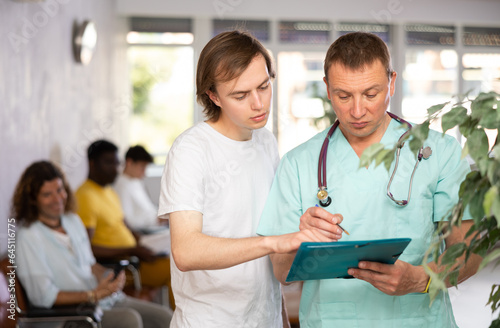 Professional doctor showing papers while explaining results of medical examination to young male patient in lobby of clinic