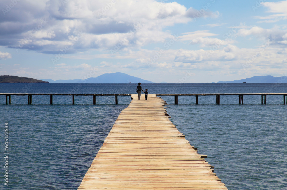 Naklejka premium Mother and son walking on a wooden pier background with blue sky and white clouds