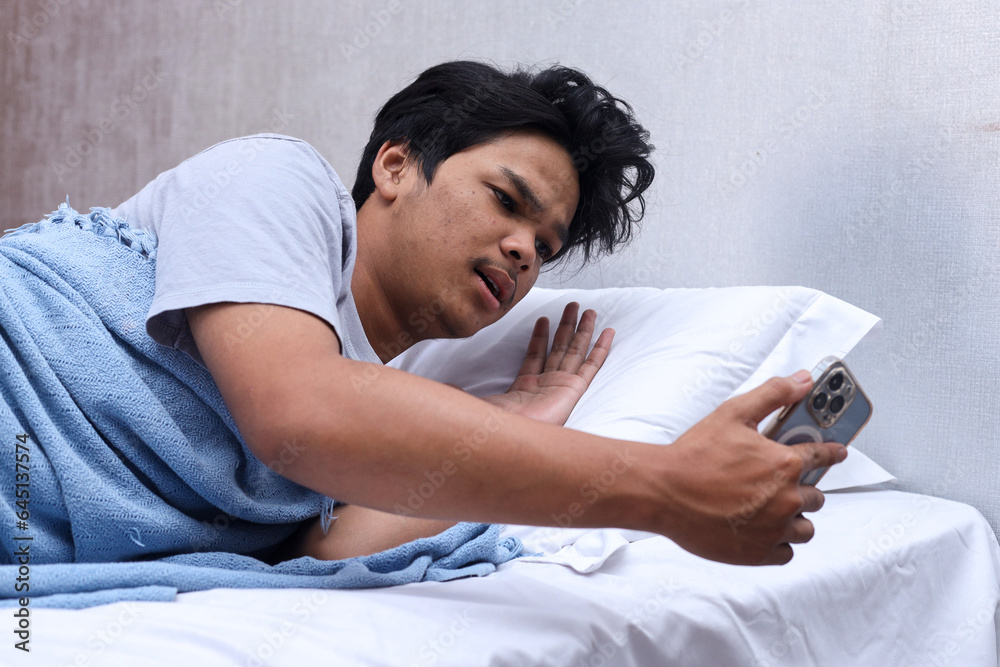Young man looking at time of alarm clock on smartphone while lying on bed in bedroom with shocked expression. Late for work.