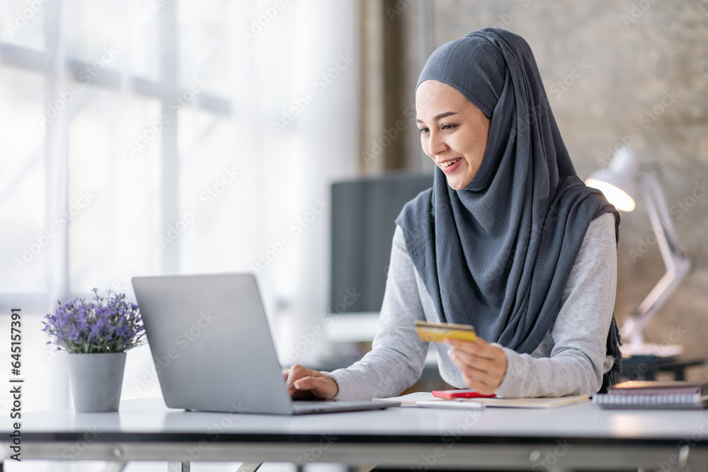 Young beautiful Asian woman in hijab sitting at home office, Muslim woman holding bank credit card and laptop, happily shopping online in online store.