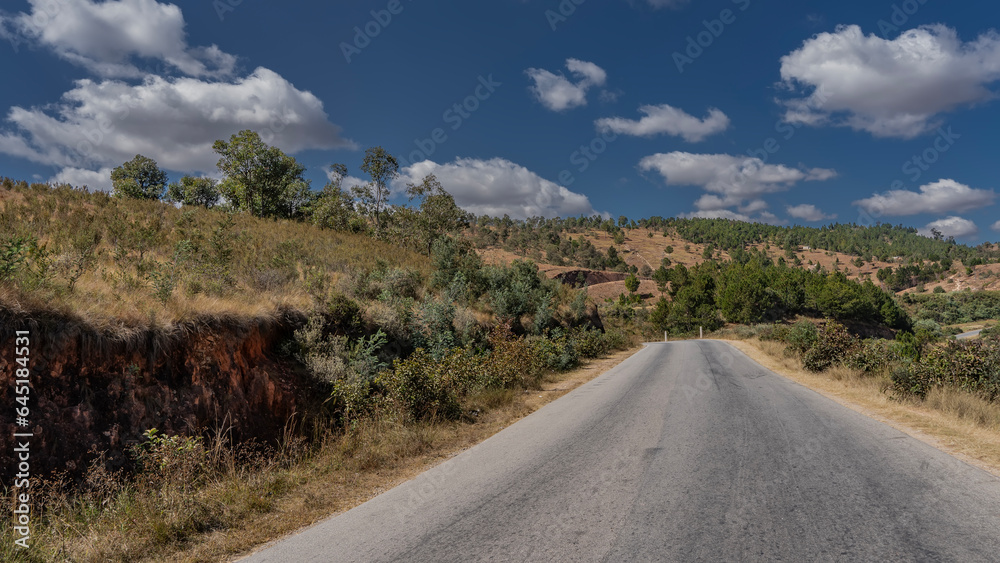A straight asphalted road goes into the distance. On the roadsides, on hills with red-brown soil, green vegetation grows. Clouds in the blue sky. Madagascar.