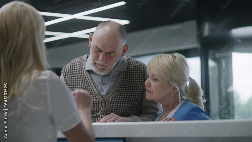 Elderly couple stand near reception desk in clinic lobby area and make appointment with doctor. Female administrator talks with patients at information counter. Medical staff work in modern hospital. Elderly couple stand near reception desk in clinic lobby area and make appointment with doctor. Female administrator talks with patients at information counter. Medical staff work in modern hospital.