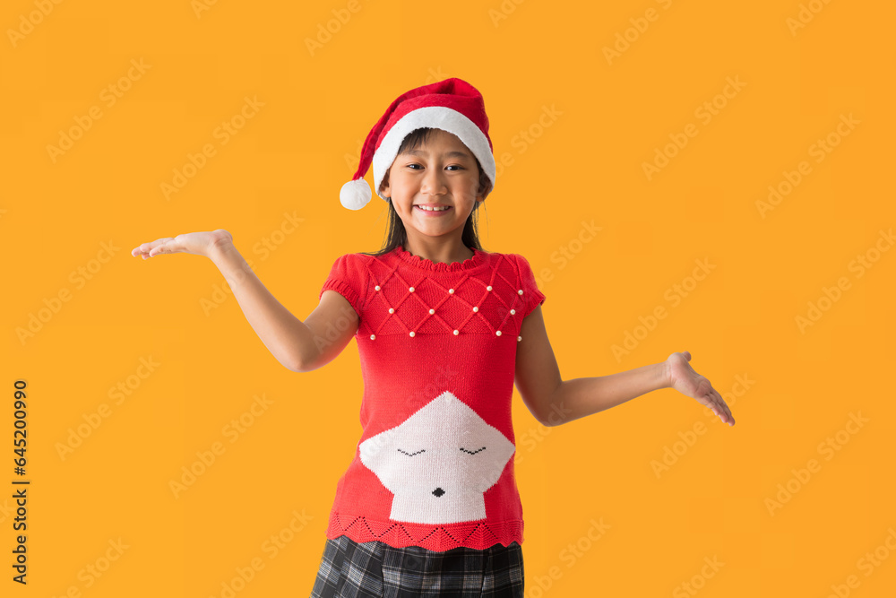 Happy smiling asian child girl in Santa Claus hat with wearing a red Christmas costume, Hold hand surprise standing posing, isolated on yellow background Happy smiling asian child girl in Santa Claus hat with wearing a red Christmas costume, Hold hand surprise standing posing, isolated on yellow background