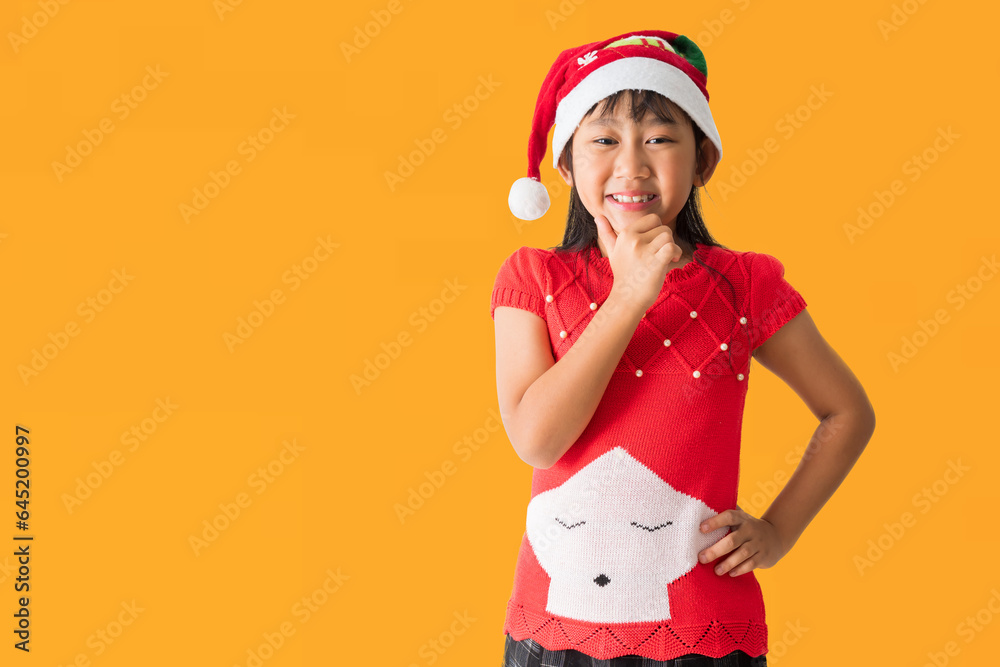 Happy smiling asian child girl in Santa Claus hat with wearing a red Christmas costume, Looking surprise standing posing, isolated on yellow background Happy smiling asian child girl in Santa Claus hat with wearing a red Christmas costume, Looking surprise standing posing, isolated on yellow background