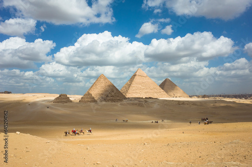 Giza pyramid complex in Egypt  The three main pyramids together with subsidiary pyramids  Tourists sightseeing on camels and horses  White clouds on blue sky  Shadow from clouds on desert yellow sand
