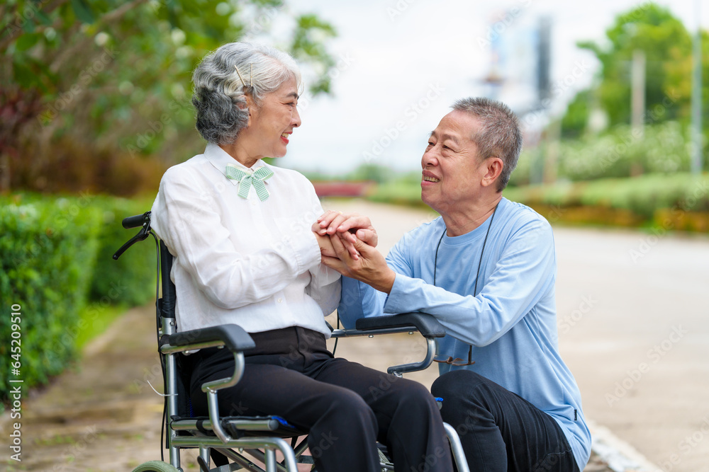 Mature asian female people sitting on wheelchair is closely monitored by her husband. elderly couple at park outdoor, relax, elderly caregiving, husband devotion to wife in park setting Mature asian female people sitting on wheelchair is closely monitored by her husband. elderly couple at park outdoor, relax, elderly caregiving, husband devotion to wife in park setting