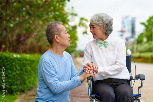 Mature asian female people sitting on wheelchair is closely monitored by her husband. elderly couple at park outdoor, relax, elderly caregiving, husband devotion to wife in park setting