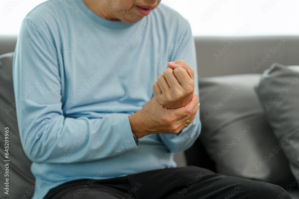 Elderly Asian gentleman with wrist discomfort on the couch, expressing pain due to Carpal Tunnel Syndrome.