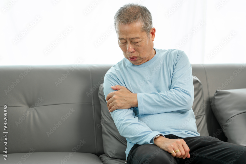 Senior Asian man suffering from intense shoulder pain on the sofa, in need of comfort. Painful moment