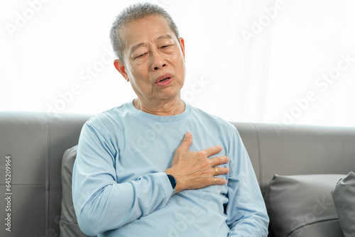 Elderly asian man on sofa, clutching chest in severe pain, showing signs of possible heart attack at home. serious health concern, home emergency, health crisis, chest pain alarm © makibestphoto