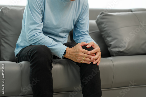 Senior elderly asian in blue shirt man suffering from intense knee pain on the sofa, seeking relief.
