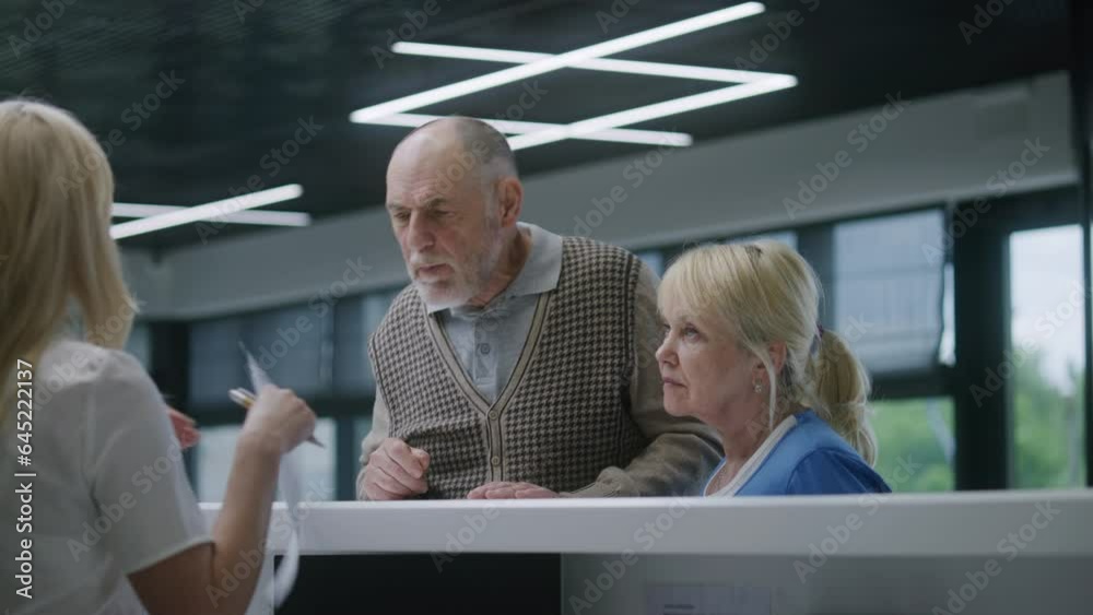 Elderly couple stand near reception desk in medical center waiting room and make appointment with doctor. Mature doctor consults patients at information counter. Medical staff work in modern clinic. Elderly couple stand near reception desk in medical center waiting room and make appointment with doctor. Mature doctor consults patients at information counter. Medical staff work in modern clinic.