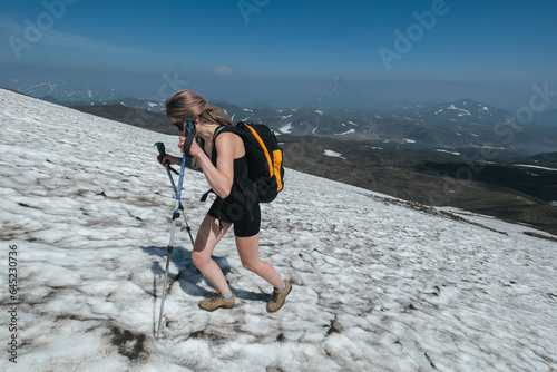 Woman hiker wearing shorts backpack and trekking poles walking on snowy slope to the top of volcano Gorely, Kamchatka photo