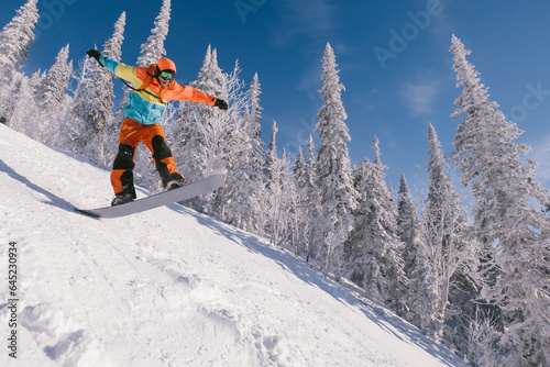 Male snowboarder wearing coloured clothing jumping in snow covered spruce trees on sunny day