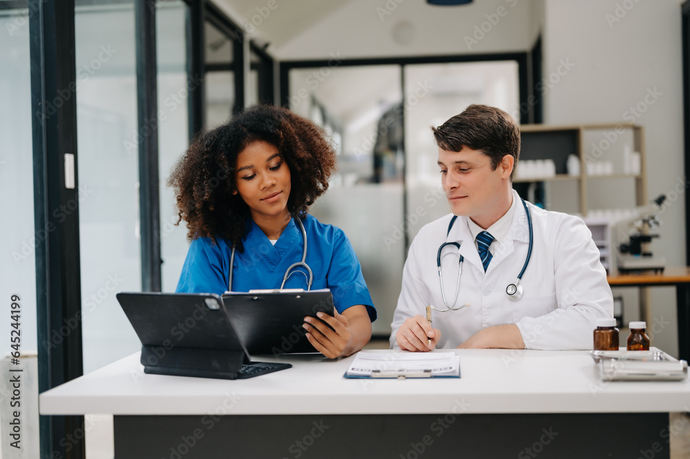 Focused mature male doctor and African female nurse look at tablet and laptop discuss anamnesis together. Concentrated diverse medical professionals in hospital. Focused mature male doctor and African female nurse look at tablet and laptop discuss anamnesis together. Concentrated diverse medical professionals in hospital.