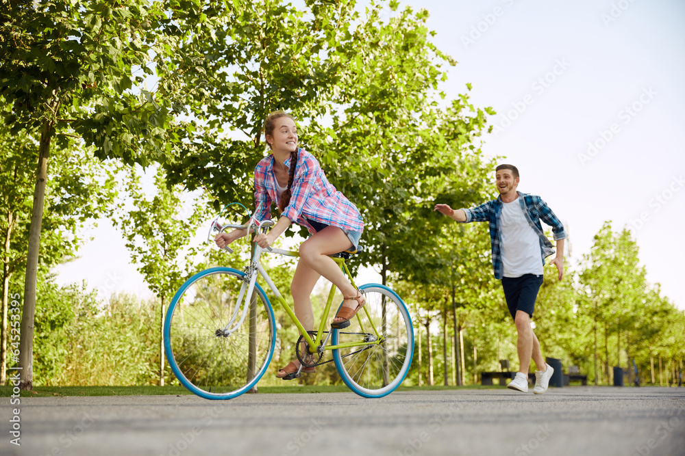Fototapeta premium Cheerful active young couple with bicycle in public park together having fun. Man catches up with girlfriend on bike. Happy couple riding cycle