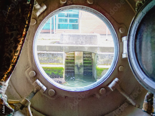 Open porthole of a very old ship looking out to a dock wall.