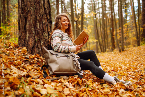 Curly-haired woman in a stylish sweater with a book sits under a maple tree in the autumn forest  reads  enjoys the weather. Concept of autumn  holiday.