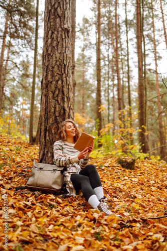 Curly-haired woman in a stylish sweater with a book sits under a maple tree in the autumn forest, reads, enjoys the weather. Concept of autumn, holiday.